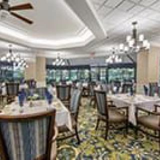 A spacious, elegantly decorated dining room with multiple tables and chairs, featuring chandeliers and large windows.