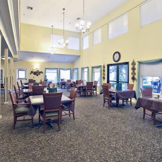 A spacious dining room with tables and chairs, illuminated by chandeliers and natural light from large windows.