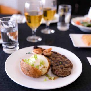 A plate of steak, shrimp, and a baked potato served with drinks on a restaurant table.