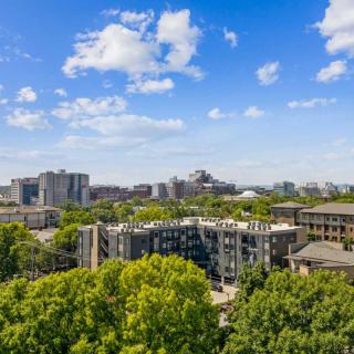 A vibrant urban cityscape with buildings surrounded by lush green trees under a bright blue sky with scattered clouds.