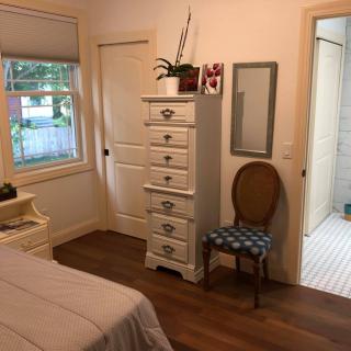 A cozy bedroom featuring a tall dresser, a wooden chair with patterned upholstery, and a mirror on the wall. A window provides natural light.
