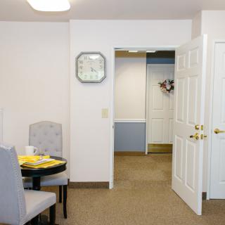 A small dining area with a table set for two, featuring an open door and a wall clock.