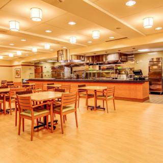 An empty restaurant dining area with wooden tables and chairs, featuring a view of the kitchen counter.