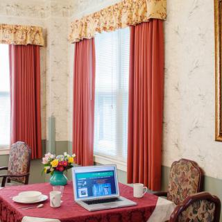 A cozy living room setup with a table, laptop, and floral arrangement near large windows with draped curtains.