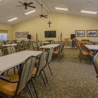 A spacious meeting room with multiple tables and chairs, featuring a cross on the wall, American flags, and a television.