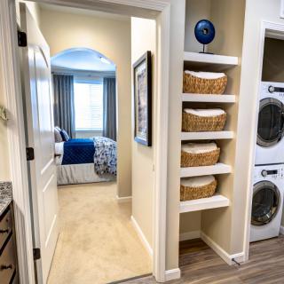 A cozy home interior featuring a hallway leading to a bedroom, with a laundry area equipped with stacked washer and dryer and shelves adorned with storage baskets.