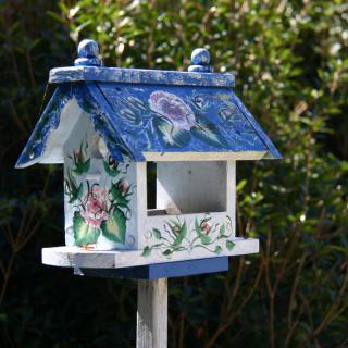 A decorative, hand-painted birdhouse with a blue roof and floral designs stands in a garden setting.