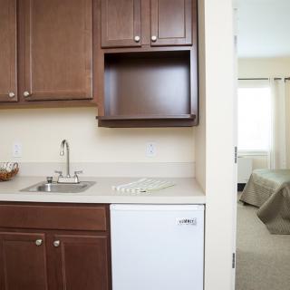 A compact kitchenette adjacent to a bedroom with wooden cabinets, a small sink, and a mini fridge.