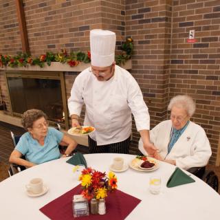 A chef serves meals to two seniors at a dining table.