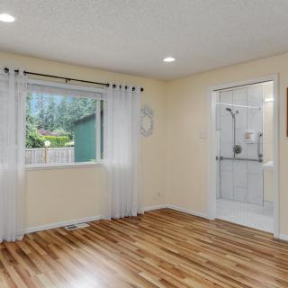 A bright bedroom with hardwood flooring, featuring a large window with sheer curtains and direct access to an adjacent bathroom.