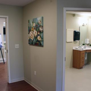 A view of a hallway in a medical facility, featuring a floral painting on the wall and an entrance to a bathroom.