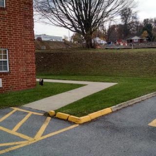 A view of a sidewalk adjacent to a brick building, leading through a grassy area with a tree in the background.