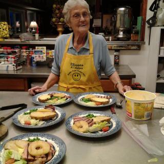 An elderly woman stands in a kitchen wearing an apron, preparing plates of sandwiches on a counter.
