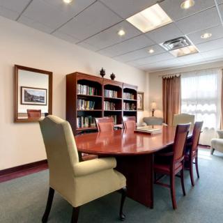 A well-lit conference room with a large wooden table surrounded by chairs, featuring a bookshelf and wall artwork.