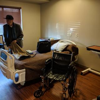 A man organizes belongings in a hospital room equipped with a bed and a wheelchair.