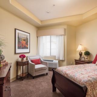 A cozy, well-decorated bedroom with a bed, chair, and a window letting in natural light.