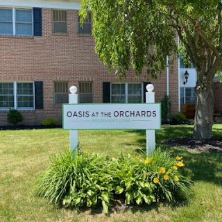 A sign for 'Oasis at the Orchards' in front of a residential building, surrounded by grass and plants.