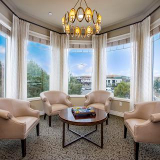 A cozy living room with armchairs arranged around a coffee table, illuminated by natural light through large windows and a central chandelier.