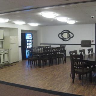 A dining area with multiple tables and chairs, adjoining a small kitchenette, featuring wooden flooring and wall art.