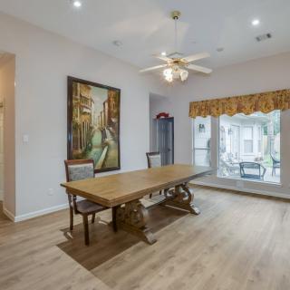 A spacious dining room with a wooden table and chairs, featuring a ceiling fan, large windows, and decorative artwork.