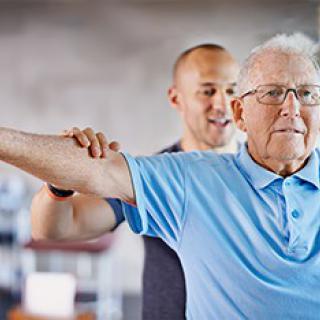 An elderly man receiving assistance from a trainer during a physical therapy session.