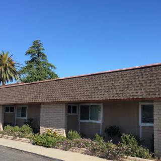 A single-story residential building with a shingled roof, surrounded by trees and landscaping under a clear blue sky.