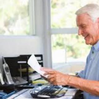 An elderly man happily working at his desk with papers and a laptop.