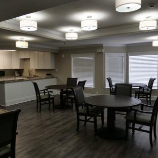 A modern dining area with round tables, chairs, and overhead lighting, adjacent to a kitchenette.