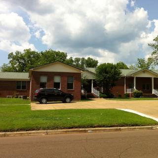 A suburban brick house with a driveway and parked car, surrounded by a well-maintained lawn and trees under a partly cloudy sky.