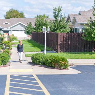 A person walking along a sidewalk in a residential neighborhood with houses, bushes, and trees.