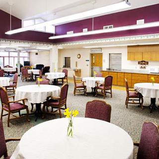 A spacious dining area with round tables covered in white tablecloths and adorned with yellow flowers, featuring a kitchen area in the background.