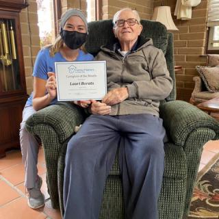 An elderly man seated in a comfortable chair next to a caregiver holding a certificate.