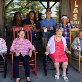 A group of elderly people and caregivers smiling while seated on rocking chairs outdoors.