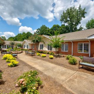 A well-maintained garden pathway lined with benches and greenery in front of a brick building under a partly cloudy sky.