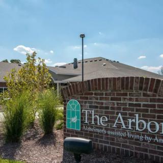 A brick sign for 'The Arbors' in front of a building on a sunny day.