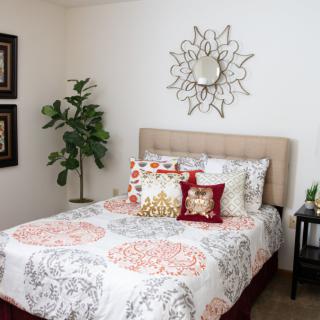 A stylish bedroom with a neatly made bed, decorative pillows, wall art, a sunburst mirror, and a potted plant.
