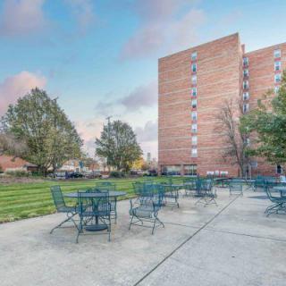 A patio with tables and chairs set outside a multi-story brick building with surrounding greenery.
