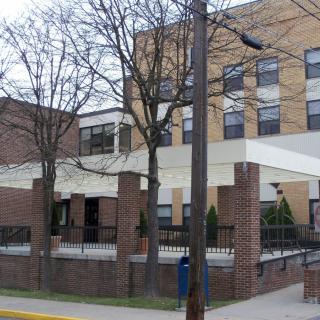 An exterior view of a brick building, likely a hospital, with trees and an entrance canopy.