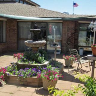 A quaint courtyard with a central fountain surrounded by colorful flowers and outdoor seating, beside a brick building displaying a flag.