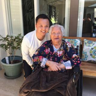A young man poses happily with an elderly woman seated in a wheelchair, surrounded by a garden setting.
