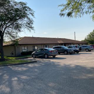 A view of a building with a parking lot filled with cars, surrounded by trees on a sunny day.