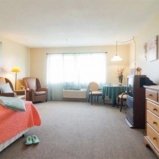 A well-lit bedroom interior with a bed, armchair, dresser, and a window allowing natural light in.