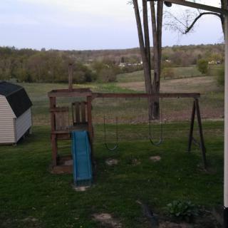 A backyard view featuring a swing set, slide, and shed with a scenic tree-filled landscape.