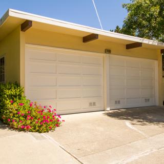 A sunny view of a residential garage with two closed doors, surrounded by greenery and flowers.
