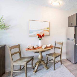 A cozy dining area featuring a small round table set for two, adorned with flowers, and accompanied by wooden chairs. A fridge stands in the corner, completing the minimalist kitchen decor.