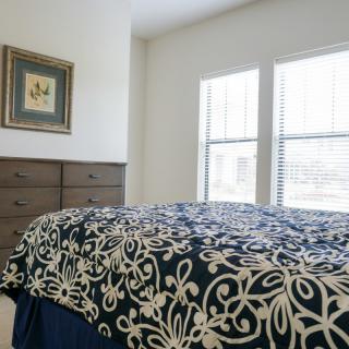 A cozy bedroom featuring a bed with patterned bedding, a wooden dresser, and large windows.