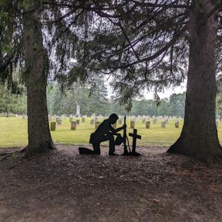 A silhouette of a person kneeling at a grave, surrounded by trees in a serene cemetery.