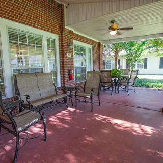 A cozy outdoor patio area with seating, tables, and a ceiling fan, surrounded by brick walls and windows.