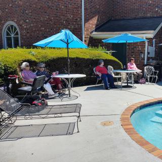 A group of people relax by the poolside under blue umbrellas on a sunny day.