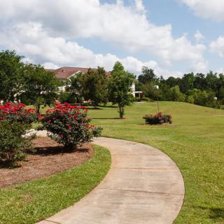 A scenic pathway winding through a landscaped park with green grass, trees, and bushes under a partly cloudy sky.